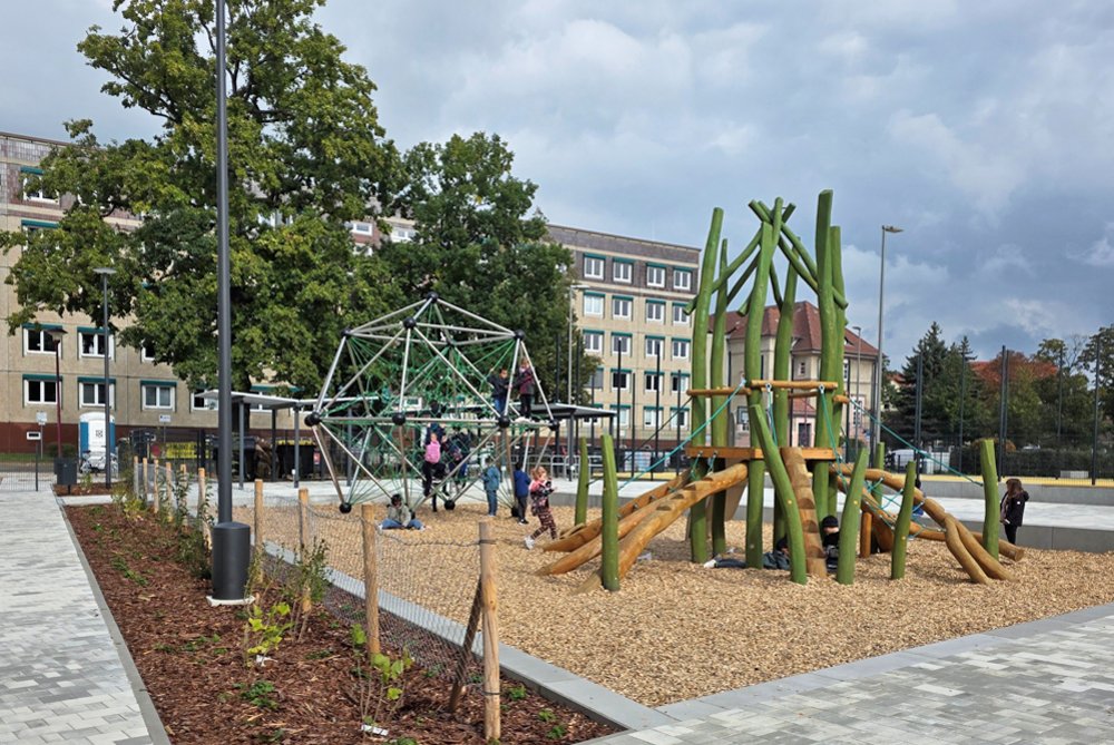 Spielplatz mit Klettergerüst aus Seilen und Holzspielgerät auf Holzschnitzelboden, umgeben von gepflastertem Weg und Gebäuden im Hintergrund