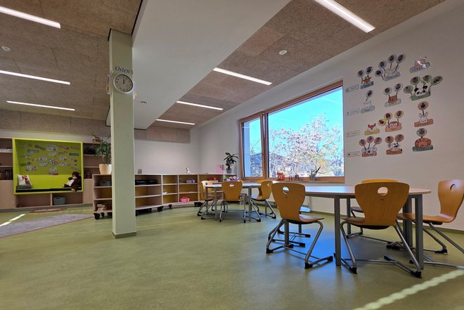 Gruppenraum mit Tischen und Stühlen vor einem großen Fenster sowie Regalen und einer Pinnwand mit Kinderzeichnungen an der Wand