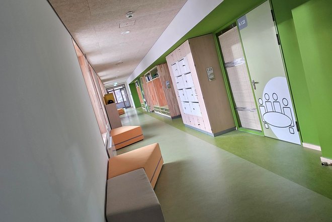 Flur mit Sitzbänken und grüner Wand mit Tür, auf der ein Piktogramm von Menschen an einem Tisch zu sehen ist