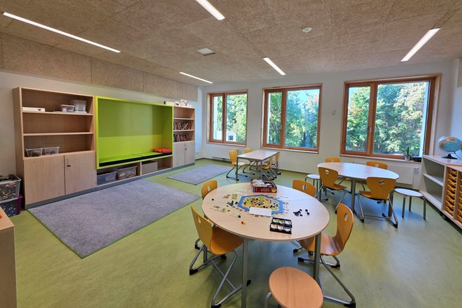Gruppenraum mit mehreren runden Tischen, gelben Stühlen, großem Teppich, Regalen und großen Fenstern mit Blick ins Grüne