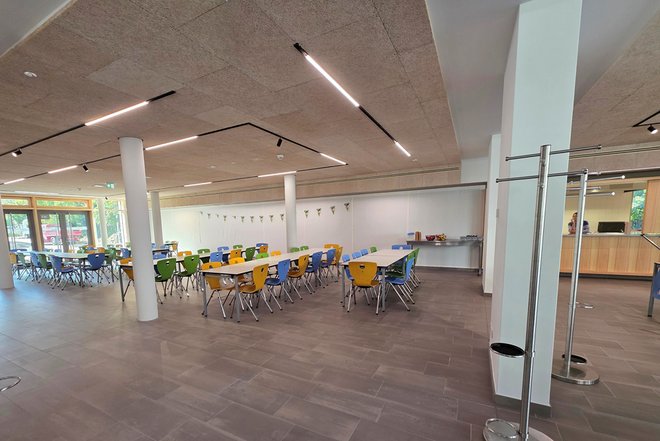 Großer heller Speisesaal mit mehreren Tischen und bunten Stühlen in Gelb, Blau und Grün, weiße Wände und große Fenster