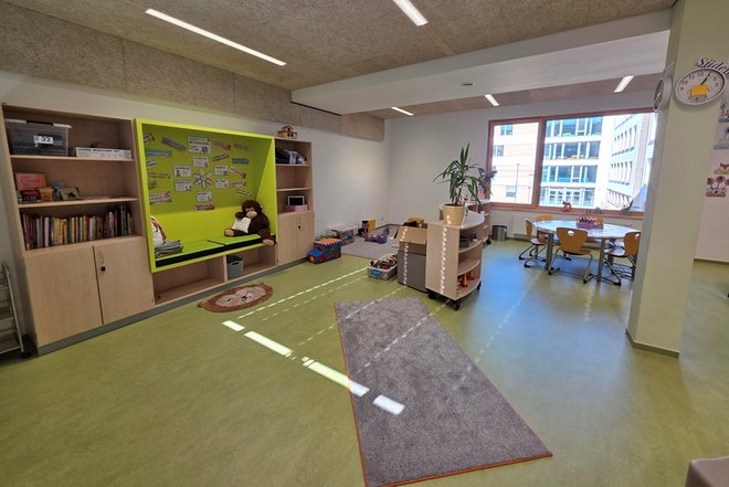 Gruppenraum mit Sitznische, Bücherregal, Spielzeug und Tischen mit Stühlen vor großen Fenstern