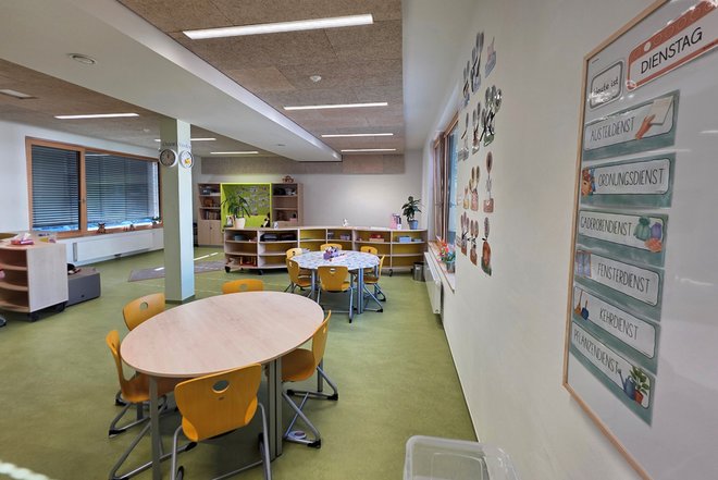 Gruppenraum mit mehreren runden Tischen und gelben Stühlen sowie einer Pinnwand mit Tagesdienstplänen an der Wand