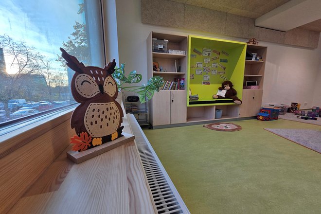 Gruppenraum mit grünem Teppich, Sitznische mit Kuscheltier, Holzregalen und Eulenfigur auf Fensterbank